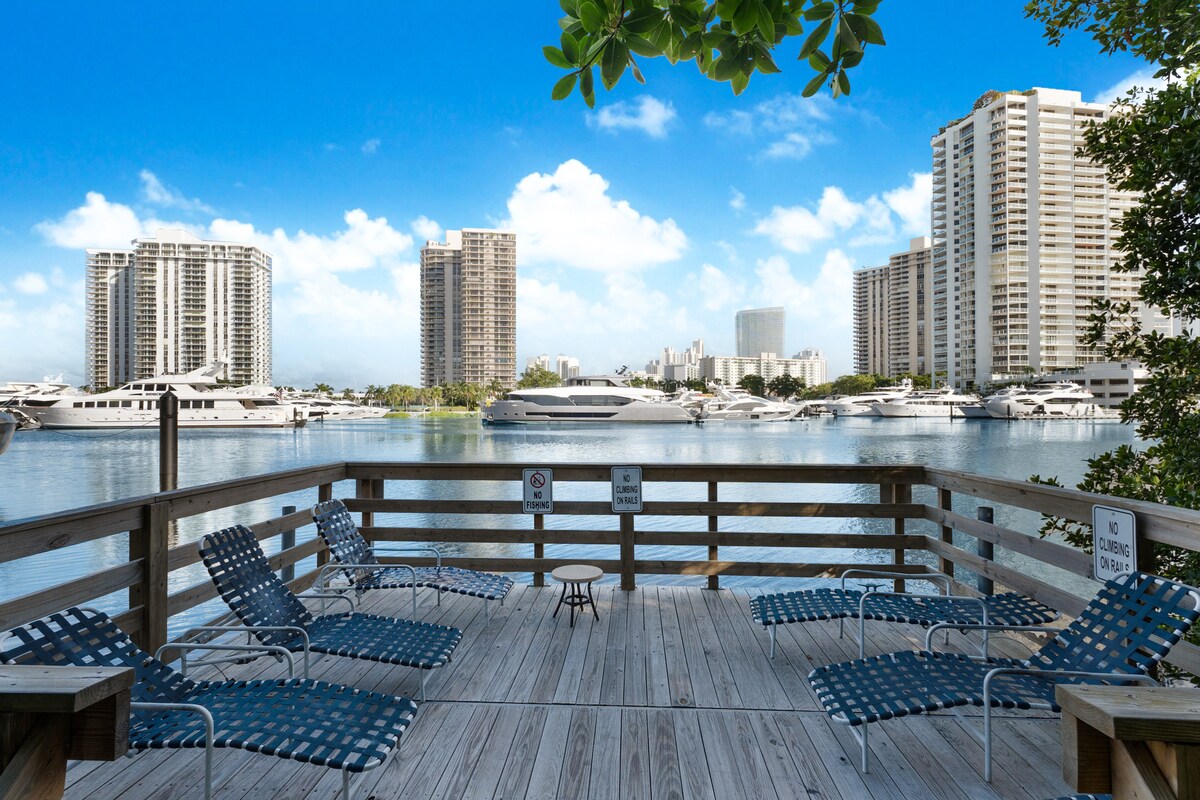 A wooden deck overlooking a calm waterway is lined with several lounge chairs. The view includes tall buildings and yachts moored nearby, framed by a clear blue sky with scattered clouds.