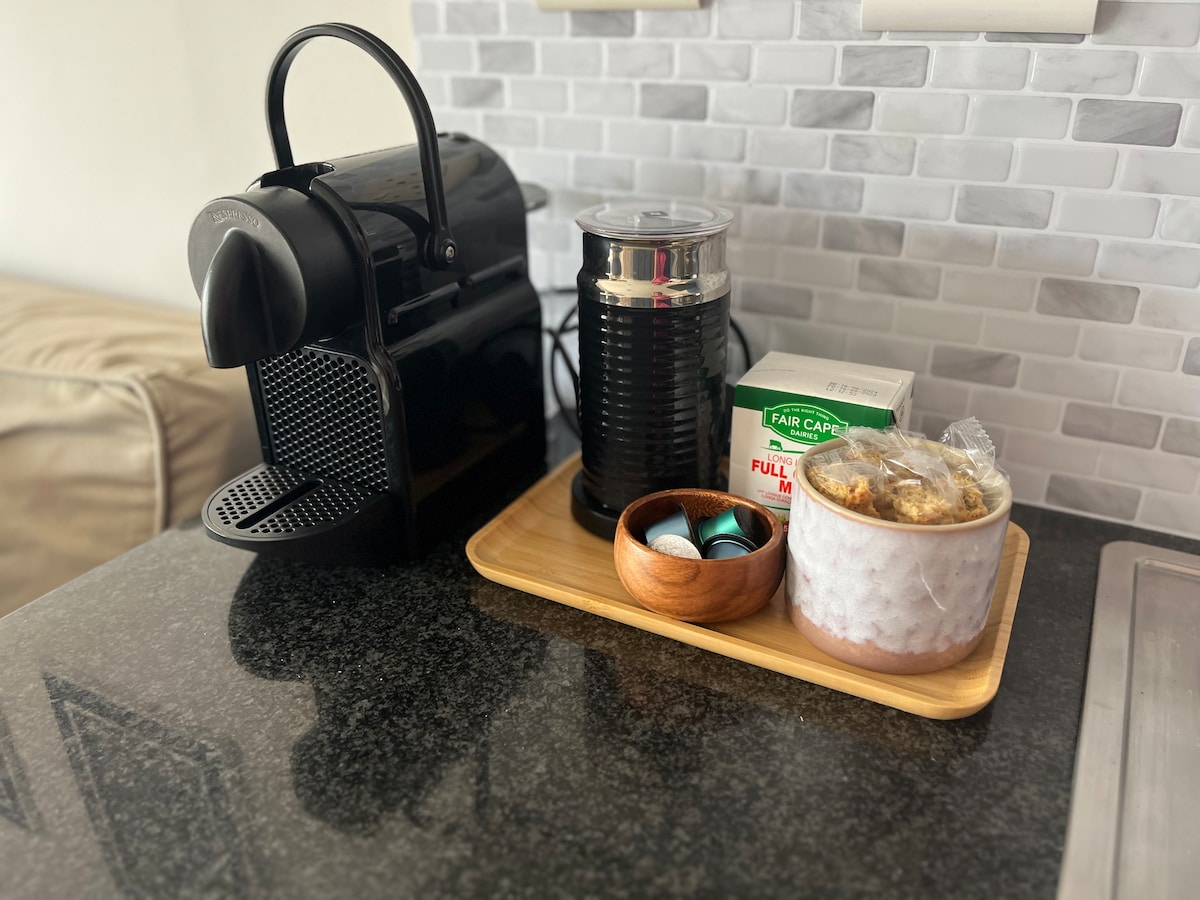 A sleek black coffee machine is positioned on a dark granite countertop, accompanied by a frother, a container of coffee pods in varied colors, and a box of milk. A wooden tray holds a small bowl of cookies, adding a touch of comfort.