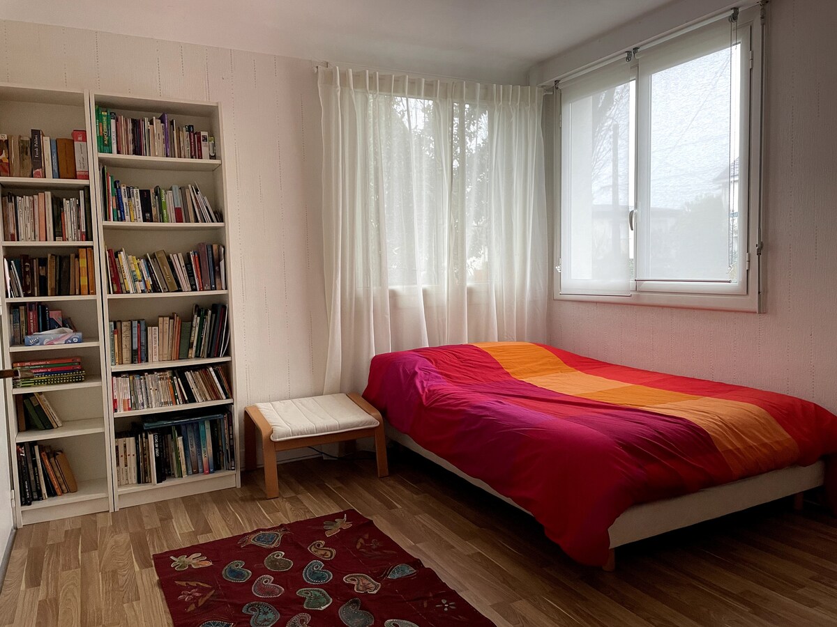 A bright bedroom is filled with natural light, featuring a spacious bed covered in a colorful orange and red blanket. A small wooden table is positioned beside the bed, and a bookshelf filled with various books stands against the wall, enhancing the cozy atmosphere.