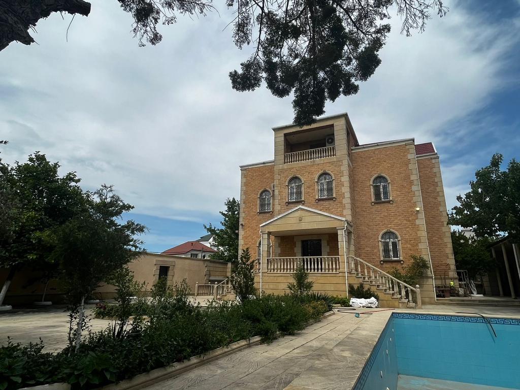The villa features a classic architectural design with a welcoming entrance and a staircase leading to a spacious balcony. Surrounding greenery adds to the charm, while a serene pool area can be seen in the foreground, inviting relaxation.