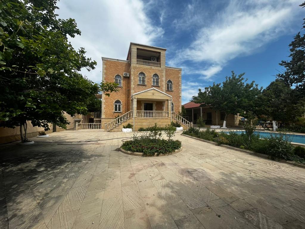 A three-story villa is showcased with a natural stone facade, surrounded by greenery and a spacious courtyard. The front features a large staircase leading to a covered balcony. A swimming pool is visible in the background, complemented by sun loungers and shaded areas.