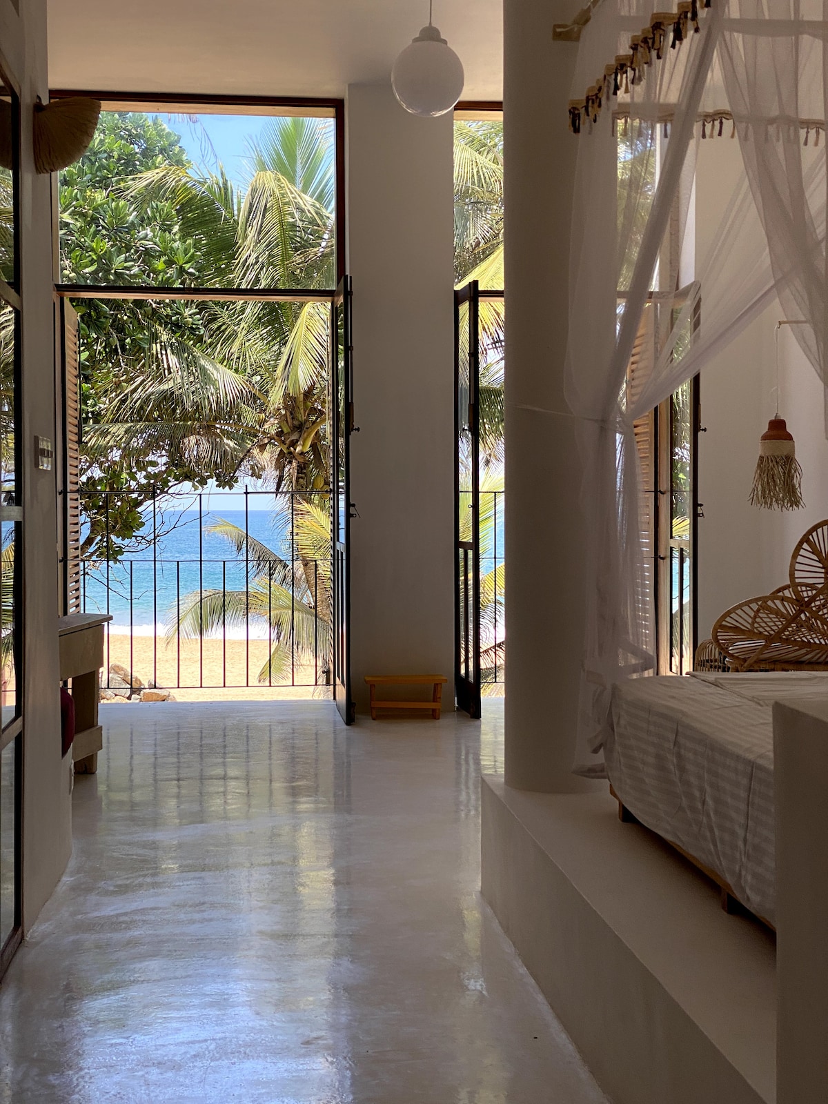 A bright and airy hallway leads to large glass doors, revealing a view of palm trees and the beach beyond. Natural light fills the space, highlighting the white walls and serene aesthetic. The scene is framed by decorative fibers hanging from the ceiling.