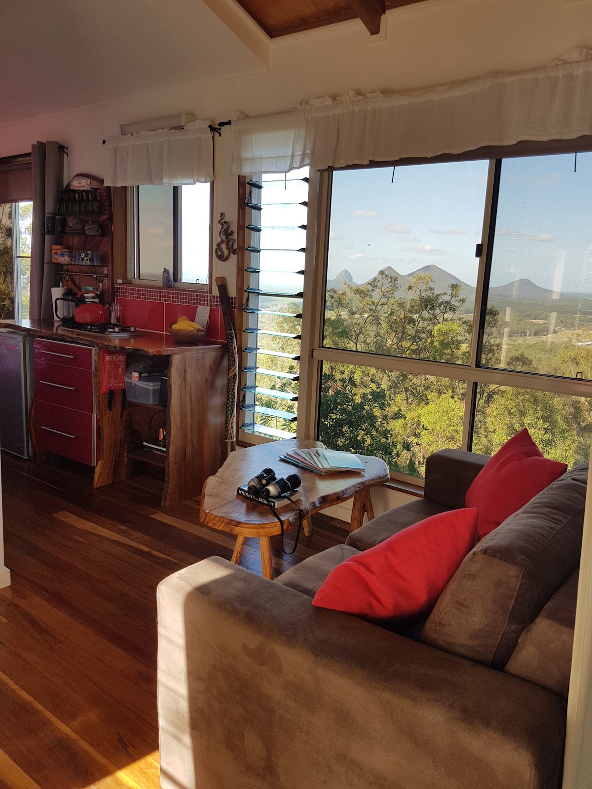 A welcoming sitting area features a comfortable gray couch adorned with red cushions, positioned beside a large window showcasing scenic views of rolling hills. A wooden table holds a camera and notebook, while a compact kitchen space with red cabinetry is visible in the background.