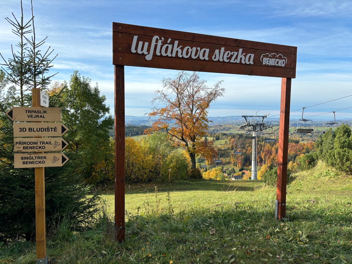 A wooden sign marks the entrance to Lufťákova stezka, with directional arrows pointing towards various trails and attractions. The sign is framed by greenery and autumnal trees, with a panoramic view of the landscape stretching in the background. A ski lift is partially visible on the right.