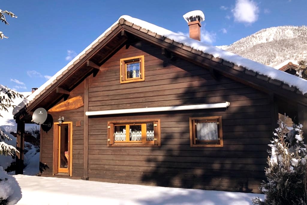 A wooden chalet features a sloping roof and charming window frames, situated amidst a snowy landscape. A satellite dish is visible on the side, and the exterior is adorned with natural wood textures, complementing the serene winter scenery.
