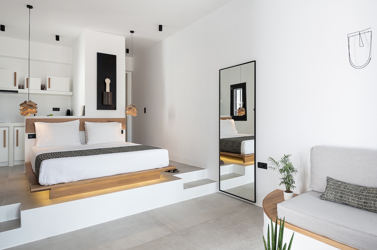 A bright and spacious bedroom is highlighted by a wooden platform bed, complemented by minimalist décor. A large mirror reflects the space, while two pendant lights add a modern touch. A comfortable seating area with a neutral sofa and a small plant is positioned nearby.