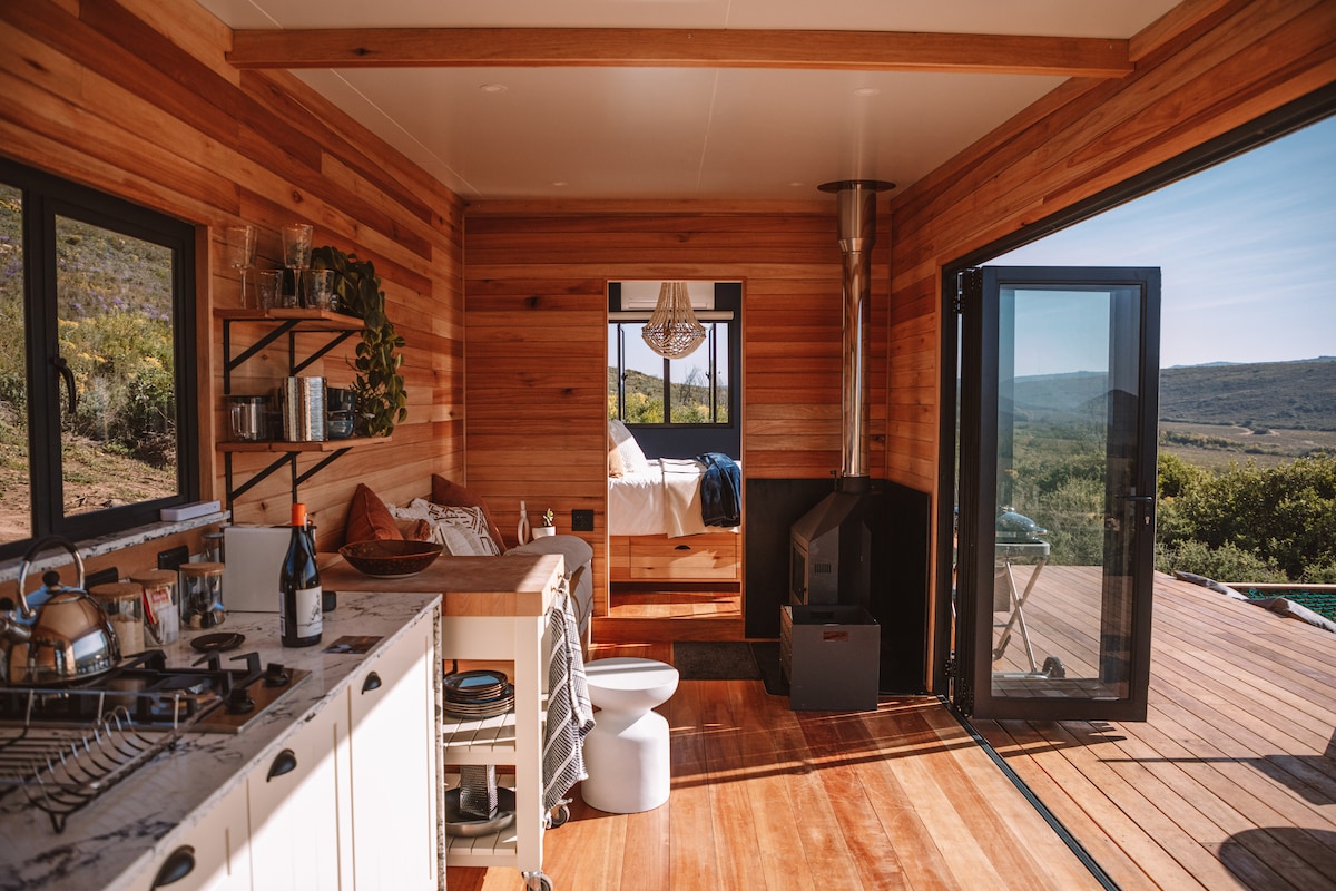 An open-plan interior showcases warm wooden paneling and large windows, allowing natural light to enhance the space. A dining area with a bar-style seating arrangement is visible, along with a well-equipped kitchen. A cozy living area is framed by a wood-burning stove, leading to an outdoor deck.