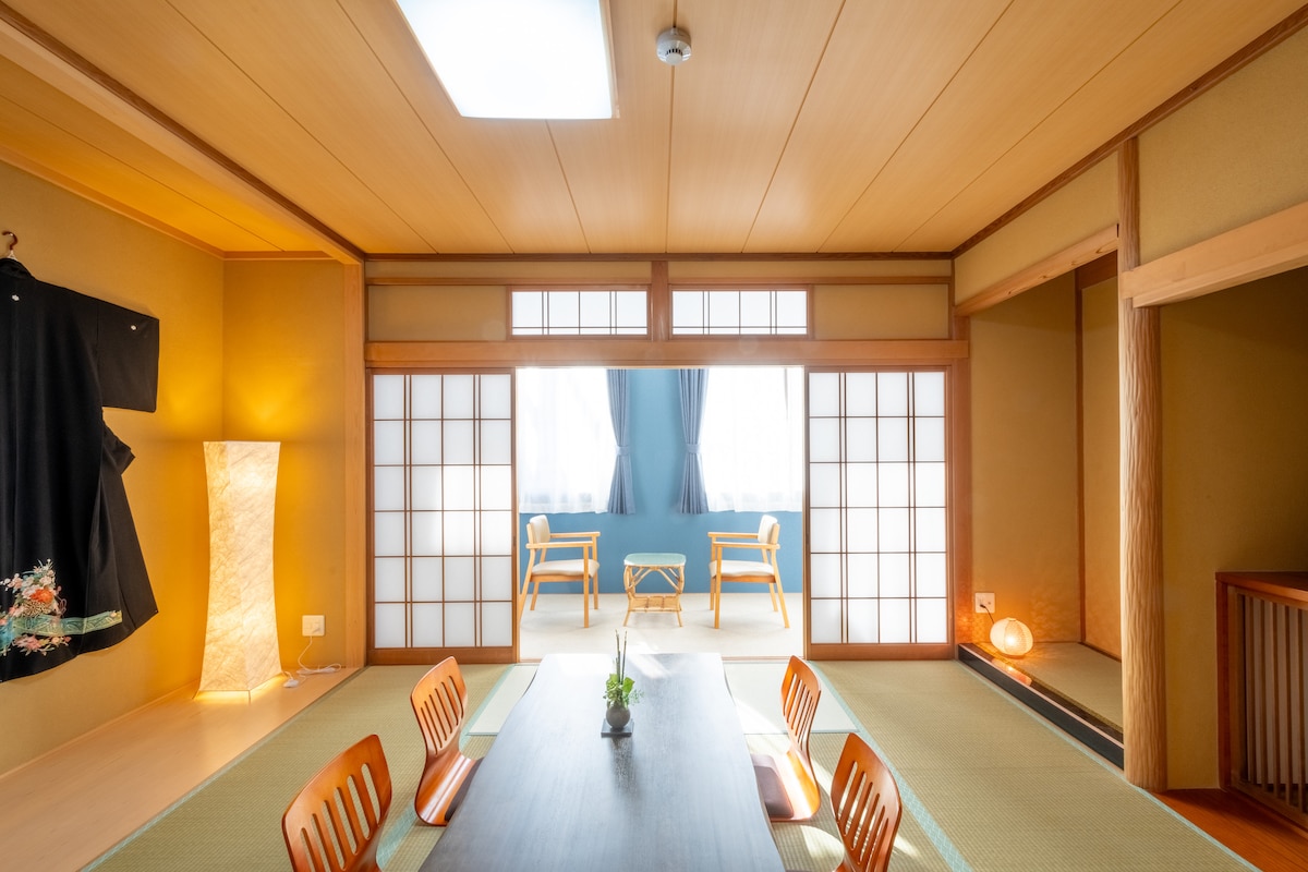 A traditional Japanese-style room features tatami flooring and a low dining table surrounded by wooden chairs. Natural light enters through shoji screens, accentuating a serene atmosphere. Two chairs are positioned near a window, creating a cozy nook for relaxation.