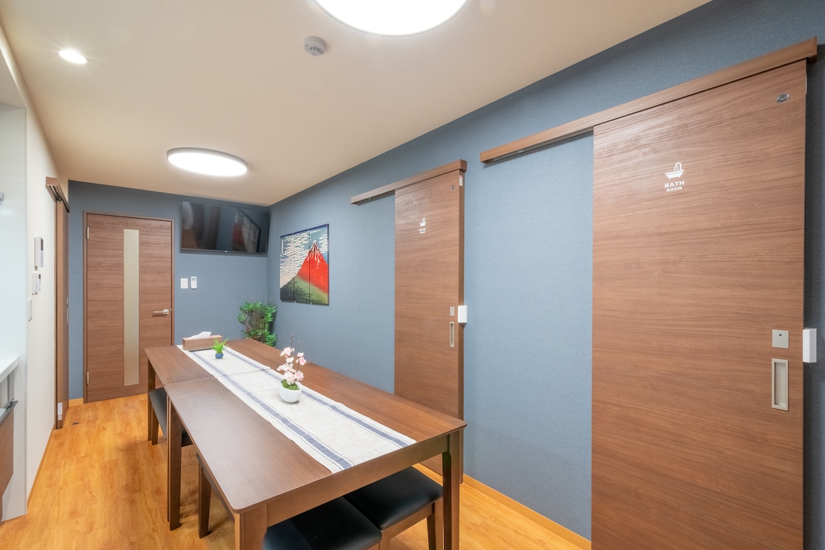 A spacious room is presented with light wooden flooring and dark blue walls. A large, rectangular dining table is centered, accompanied by dark chairs. Two sliding wooden doors are visible, along with framed artwork depicting a mountain scene on the wall.