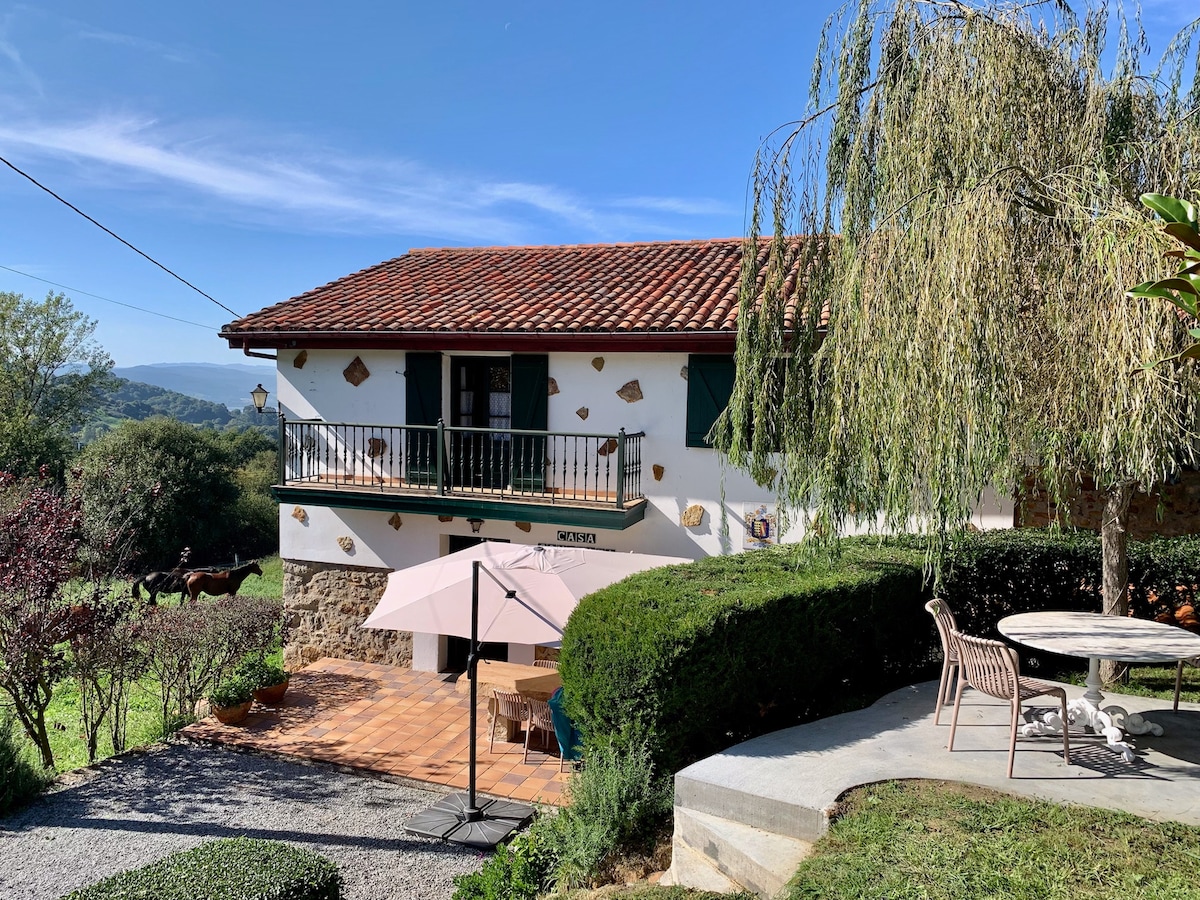 The traditional Cantabrian house is surrounded by lush greenery and features a charming patio area with outdoor seating. A large umbrella provides shade, while a round table invites leisurely gatherings. The scenic landscape and clear blue sky enhance the serene ambiance of the outdoor space.
