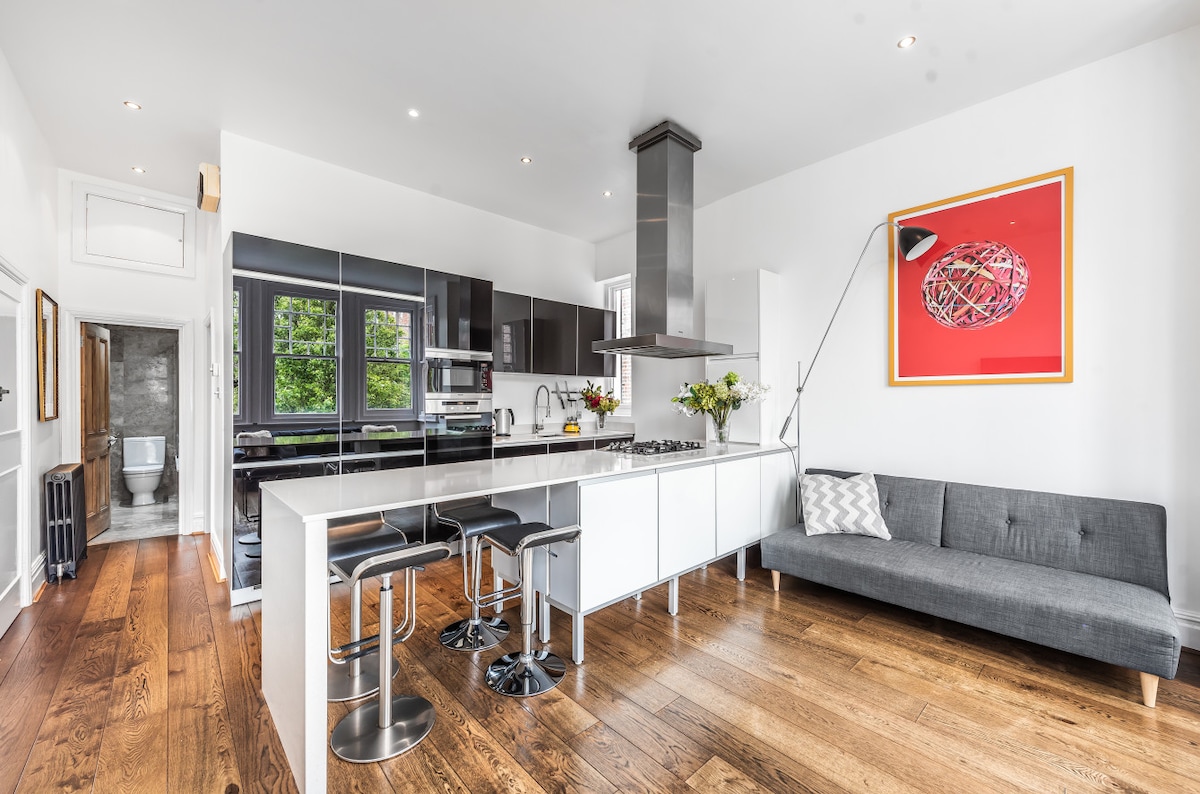 A modern kitchen is presented, featuring sleek black and white cabinetry with a large island. High-end appliances are integrated within, complemented by polished wooden flooring. A cozy gray sofa is located nearby, while a vibrant piece of wall art adds a splash of color to the space.