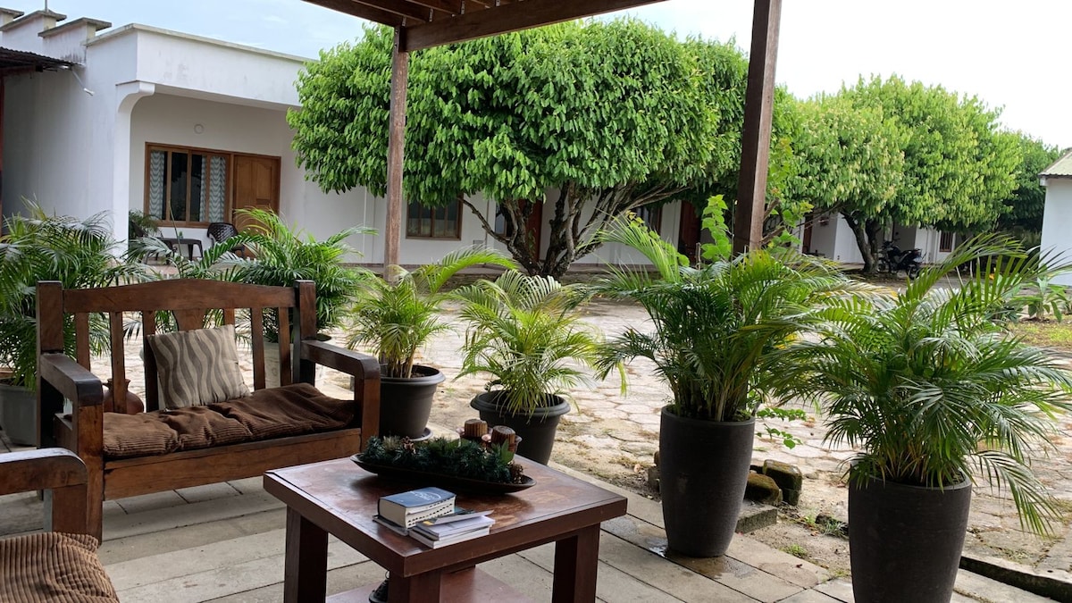 A comfortable outdoor seating area features a wooden bench with cushions and a table. Potted plants surround the space, and a pathway leads to spacious greenery beyond, creating a serene and inviting environment.