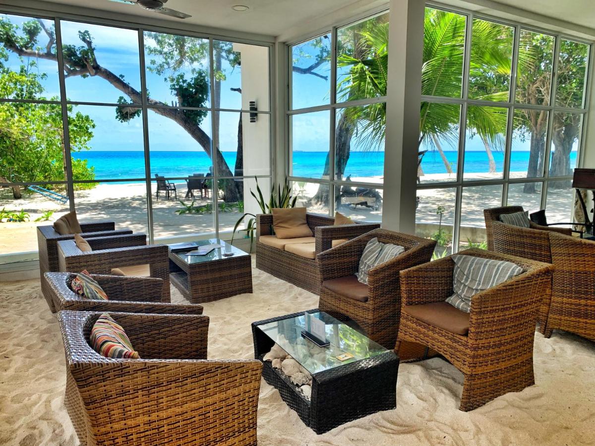 A comfortable lounge area is surrounded by large windows, offering views of the beach and ocean. Dark wicker seating is arranged around a glass-top table, with a sandy floor enhancing the tropical feel. Lush greenery is visible outside, contributing to a calming environment.