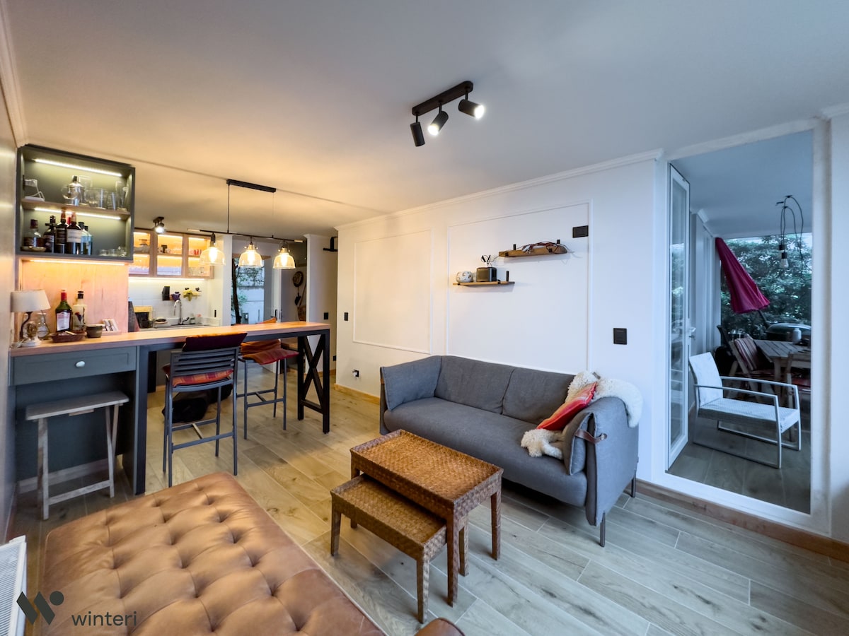 An open living area showcases a modern kitchen bar equipped with pendant lighting, adjacent to a relaxed seating arrangement featuring a sofa and coffee tables. Large windows allow natural light to flood the space, with a glimpse of the outdoor patio visible in the background.