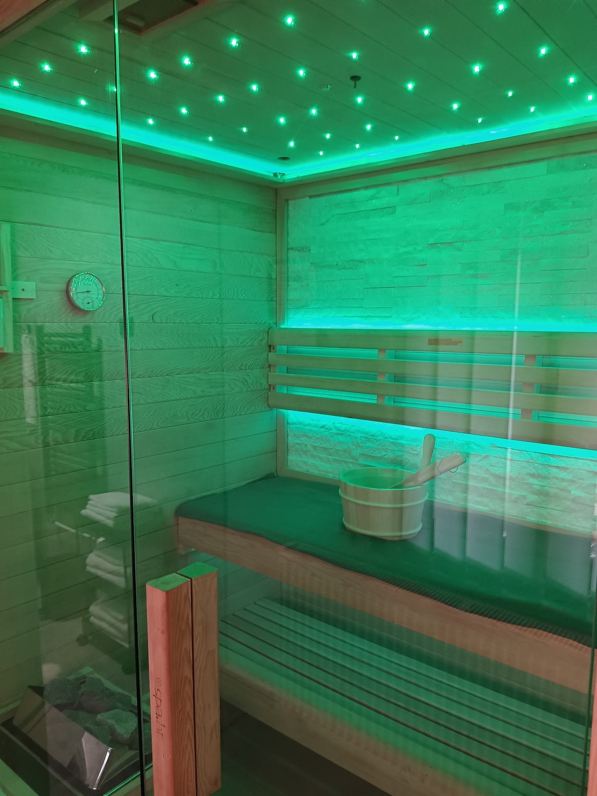 A modern sauna is showcased with a glass enclosure, accentuated by green ambient lighting. The wooden bench offers a cozy seating area. A bucket and ladle sit ready for use, contributing to the wellness experience.
