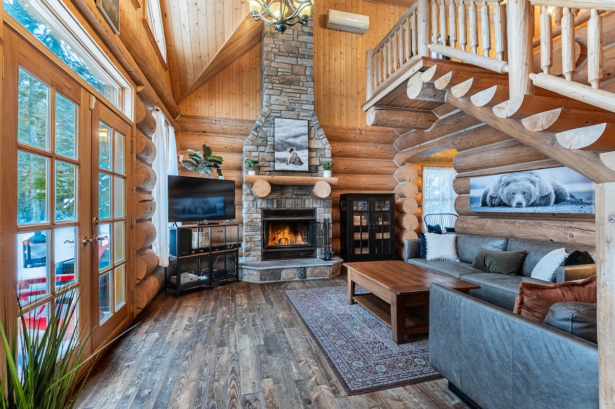A rustic living area features wooden beams and a stone fireplace as its focal point. Large windows provide natural light, while a comfortable sofa and coffee table create an inviting space for relaxation. A television is visible alongside a dark wood shelving unit, enhancing the room's functionality.