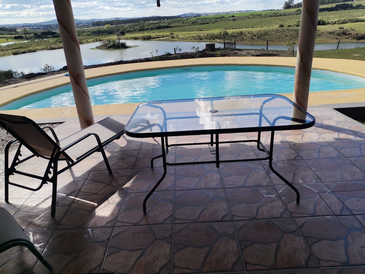 A glass-top table is positioned beside a lounge chair on a tiled patio overlooking a tranquil pool. The pool reflects the sky, and lush greenery with distant water views can be seen in the background.