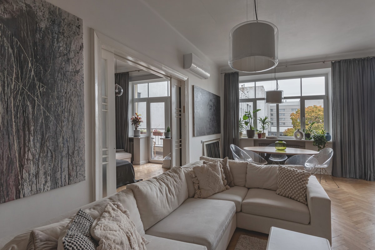 A spacious living area features a large sectional sofa with soft cushions, complemented by a variety of artworks on the walls. Natural light enters through expansive windows, illuminating a dining space adorned with plants and a round table set for meals.