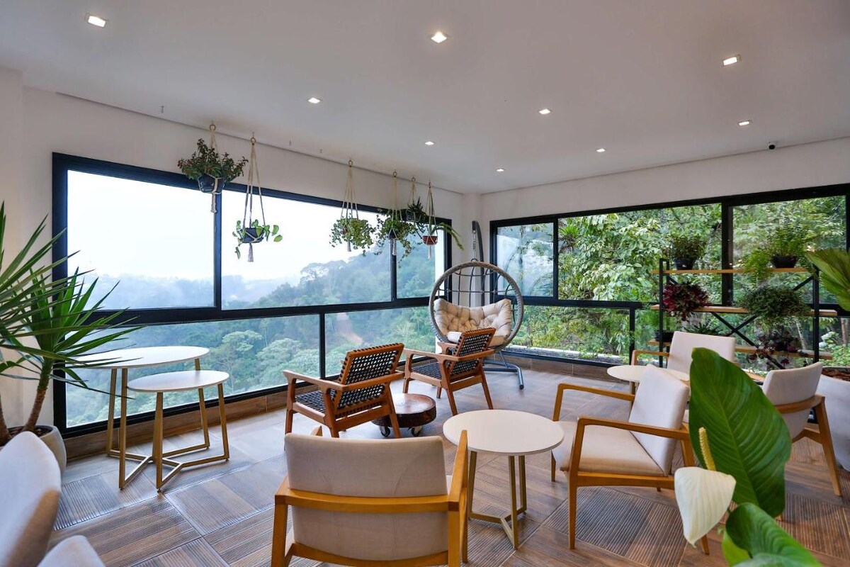 A spacious lounge area features large windows with panoramic views of the surrounding greenery. Stylish seating arrangements include comfortable chairs and small tables. Hanging plants add a touch of nature, enhancing the inviting atmosphere of the space.