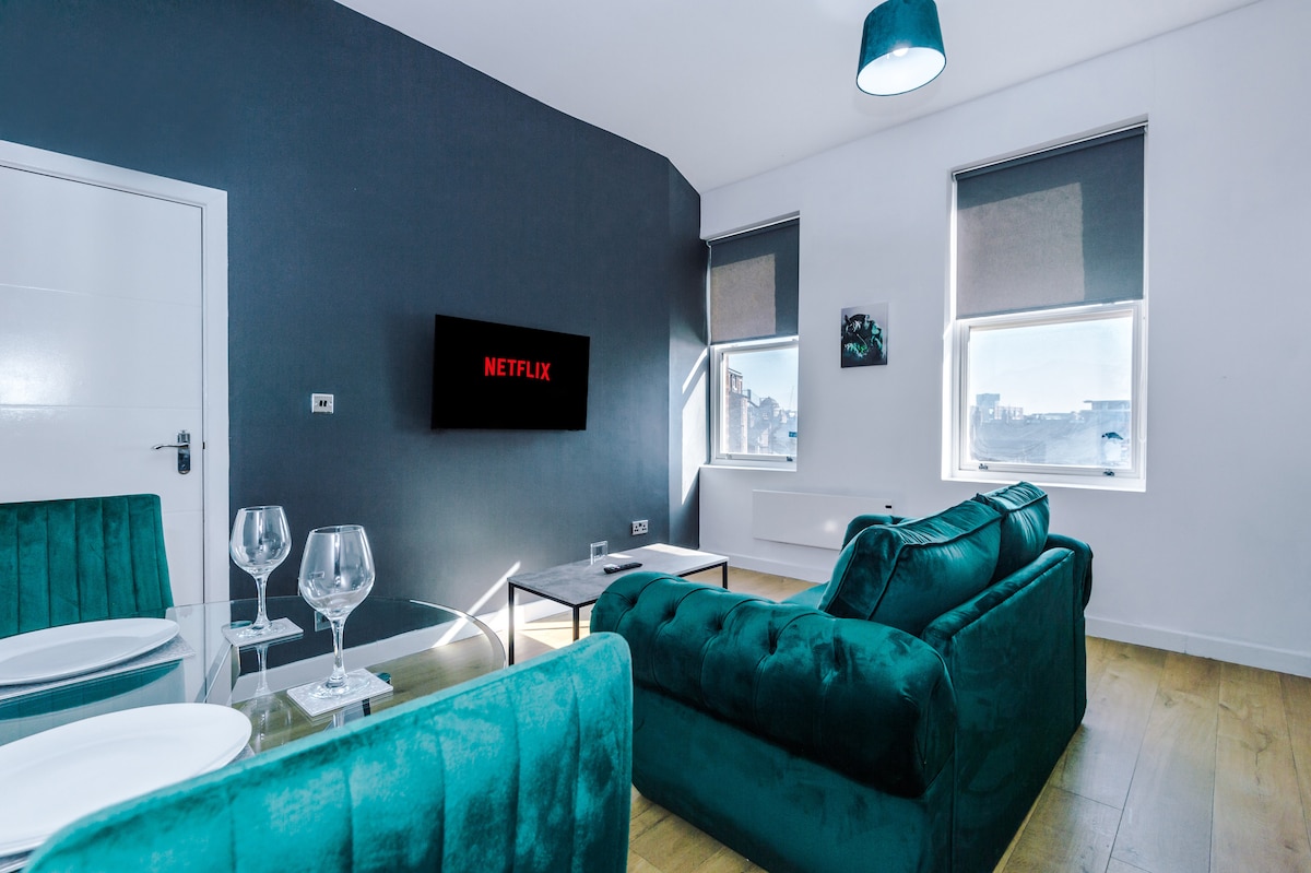 An open-plan lounge area features a plush green sofa positioned in front of a wall-mounted television displaying the Netflix logo. Two windows provide natural light, complemented by contemporary window coverings, and a dining table is set with glasses and plates.