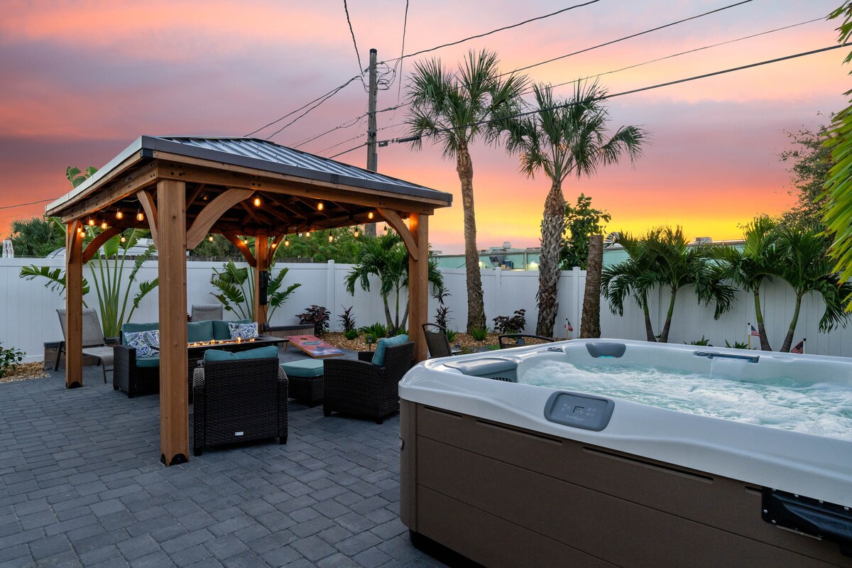 An outdoor space features a large Jacuzzi surrounded by lush greenery. A lighted gazebo with seating provides a cozy area, enhanced by the warm colors of the sunset in the background. The patio is laid with stone pavers, creating a welcoming atmosphere.