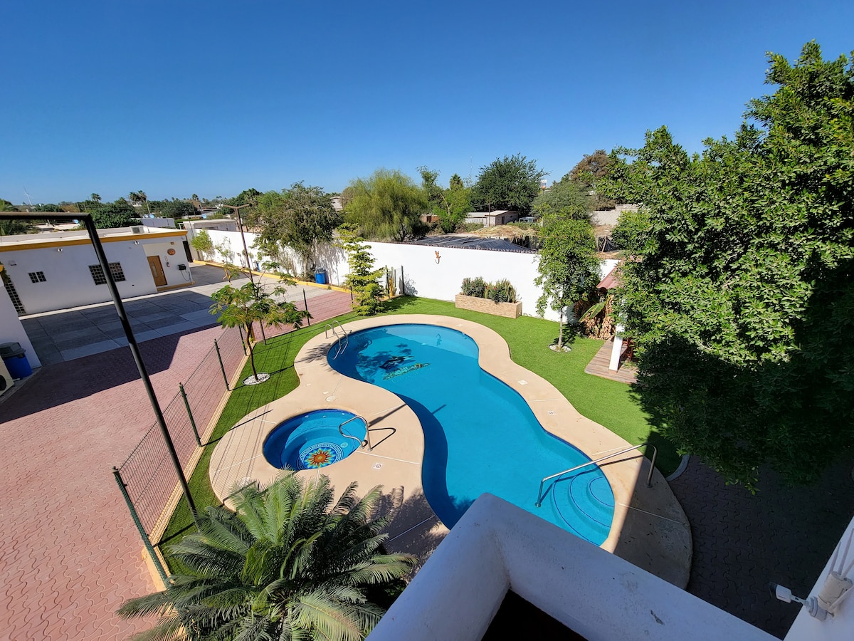 An aerial view reveals a spacious swimming pool surrounded by manicured grass and palm trees. The pool's shape is unique, with a decorative feature at its center. A patio area with lounge chairs is visible, complemented by a well-maintained garden and a nearby parking area.