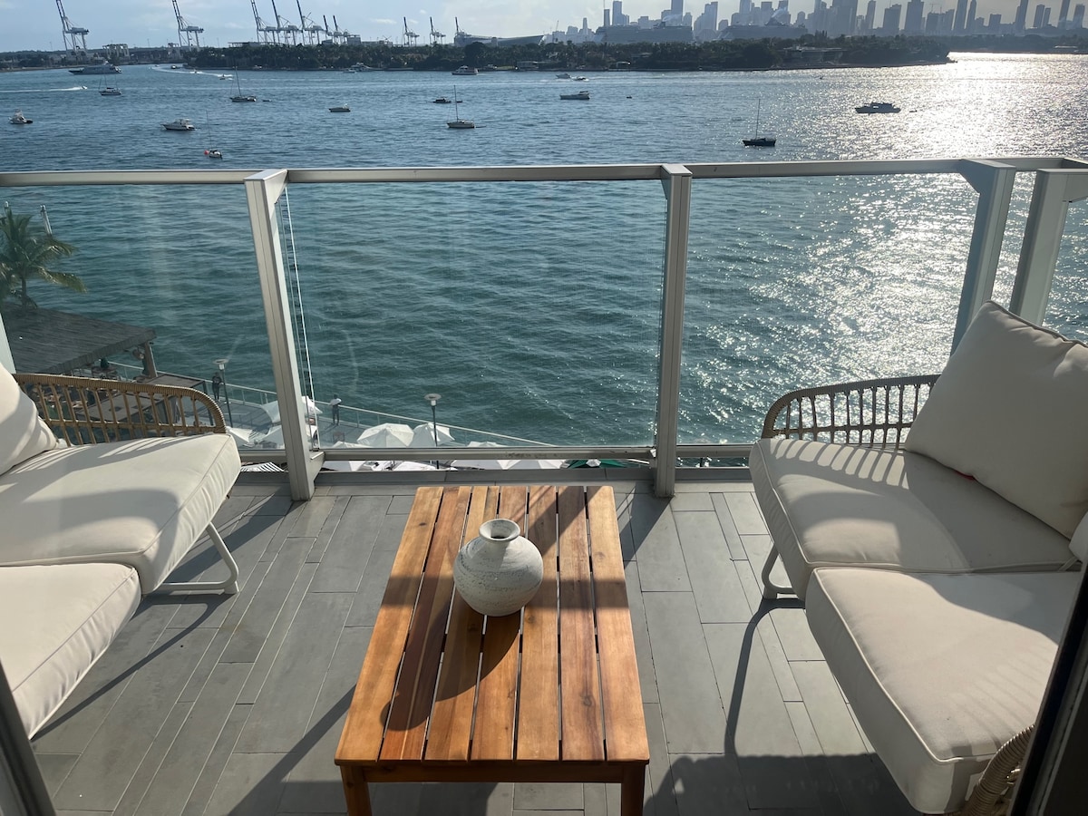 An outdoor balcony features two comfortable seating areas with cream-colored cushions. A wooden table with a decorative vase sits in the center, overlooking a view of Biscayne Bay and city skyline. The water reflects the sunlight, enhancing the serene atmosphere.