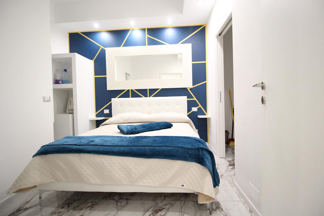 A modern bedroom features a spacious bed dressed in cream linens, complemented by a soft blue throw. A striking blue and gold accent wall adds visual interest, while a large mirror reflects natural light. Storage options are visible beside the bed.