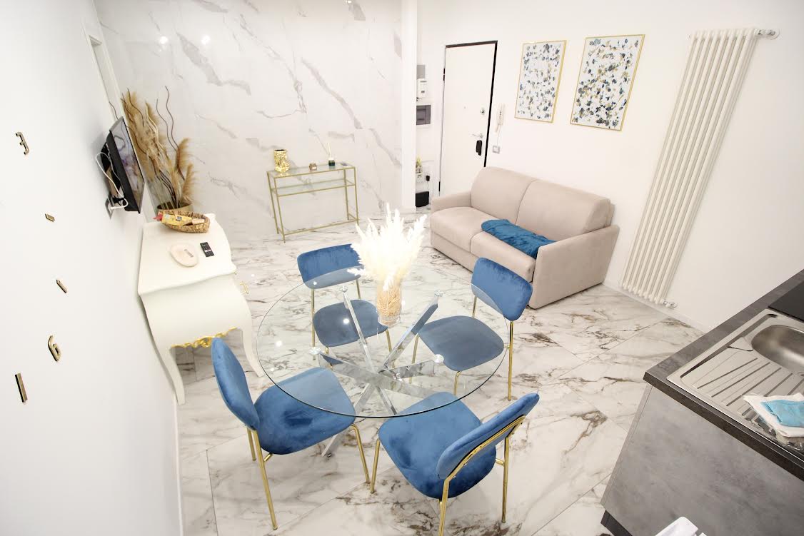 A bright living area features a round glass dining table surrounded by four blue chairs. A modern sofa sits against the wall, complemented by decorative wall art and neutral colors. The floor is adorned with marble tiles, enhancing the light and open atmosphere.