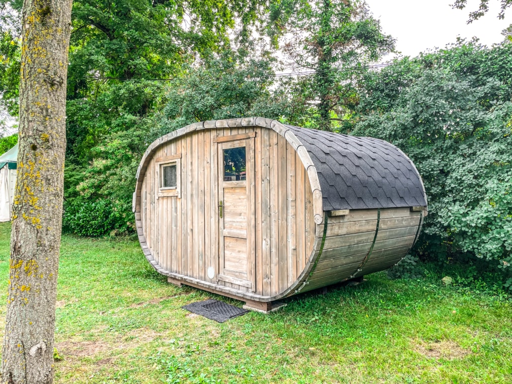 A uniquely shaped wooden cabin is nestled amidst greenery, featuring a rounded design with a sleek black roof. A small window and a door are visible on the front facade, surrounded by grass and shrubs, creating a tranquil and natural setting.