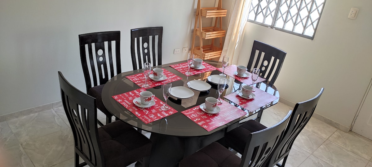 A round dining table is set for six, featuring elegant place settings with cups and plates. Dark wooden chairs encircle the table, and a tall wooden shelf is positioned in the corner. Natural light filters through a window with a patterned metal grille.