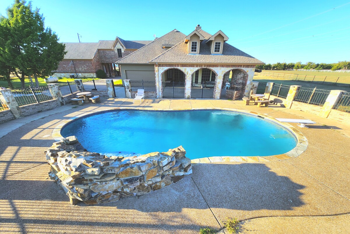 An inviting outdoor space features a well-maintained swimming pool surrounded by a stone border. Nearby, lounge chairs and a small table are positioned on the patio, offering a place to relax, while a spacious backyard extends beyond the fence.