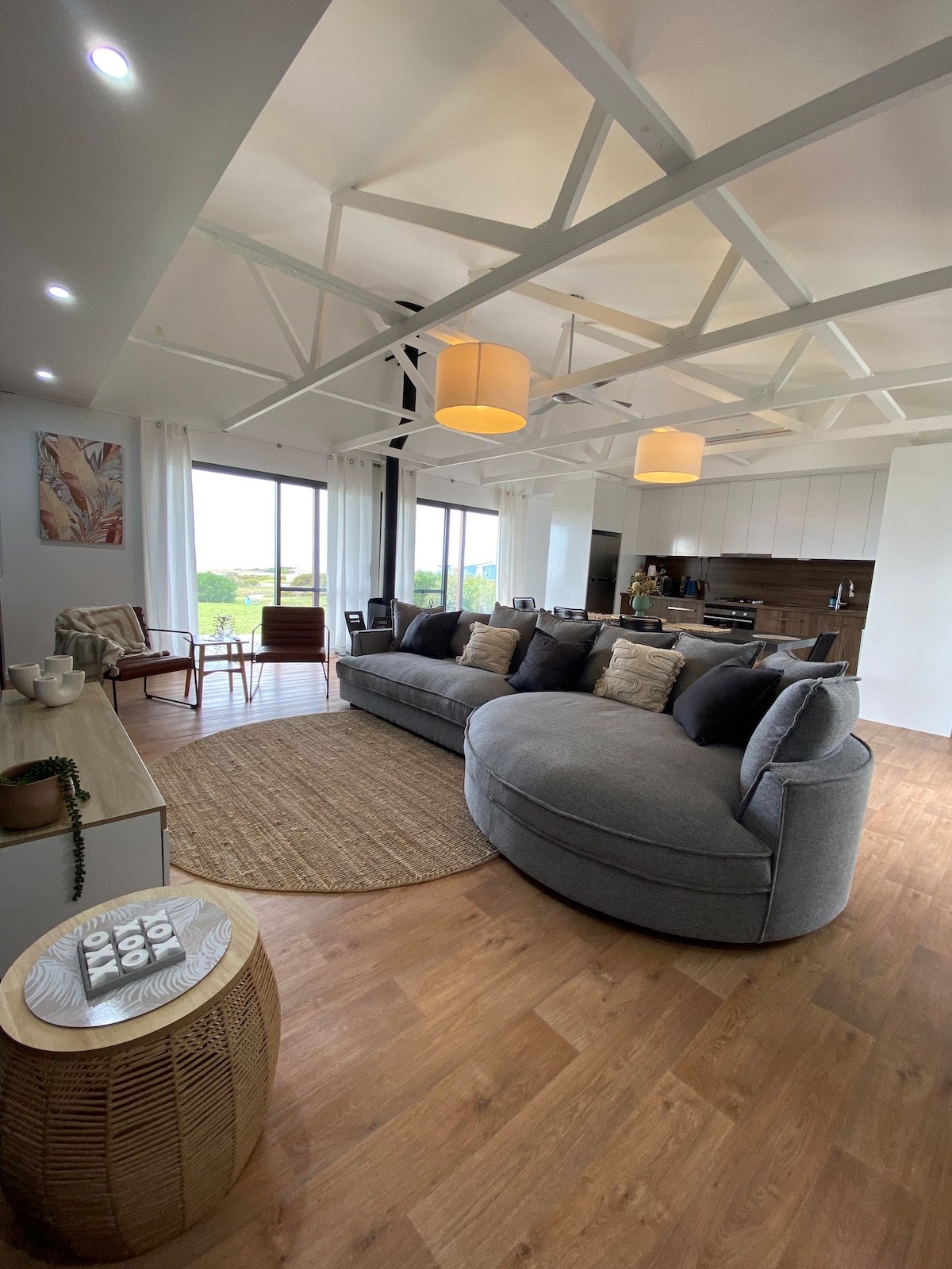 The spacious living area showcases a curved gray sofa adorned with decorative cushions, positioned on a woven rug. Large windows offer views of the exterior landscape, while natural light fills the room, complemented by contemporary light fixtures.