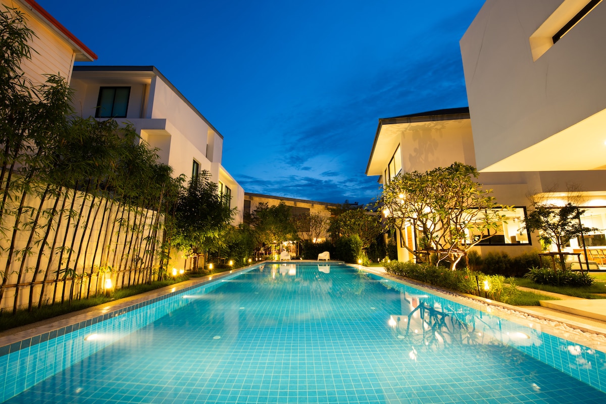 The outdoor area features a long salt-water pool, surrounded by lush greenery and softly illuminated by garden lights. The surrounding villas are partially visible, offering a serene view under the evening sky.