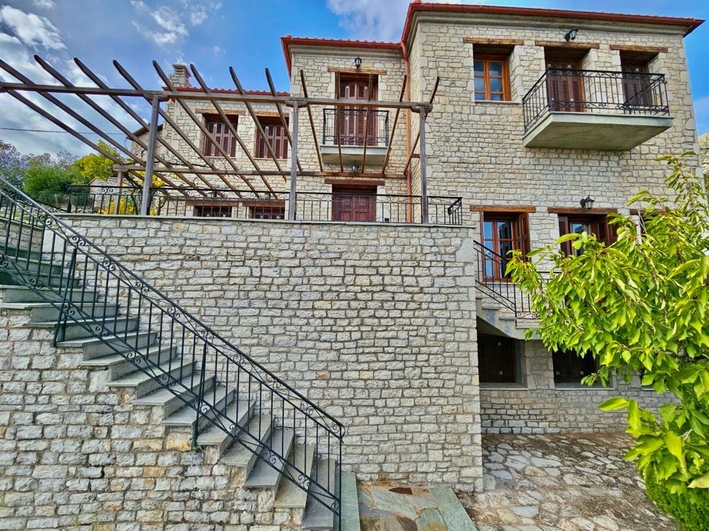A multi-level stone villa is displayed, featuring a charming exterior with wooden balconies and a partial pergola above. Stairs lead from a stone pathway into the lower level. Lush greenery frames the structure, while a clear sky provides a serene backdrop.