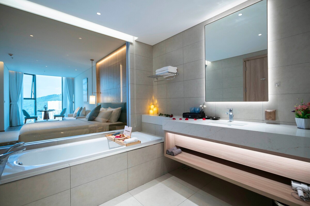 A modern bathroom features a spacious bathtub and a sleek countertop with essential amenities. A large mirror reflects the light from the room beyond, which includes a comfortable sleeping area. Natural light enters through expansive windows, enhancing the overall ambiance.