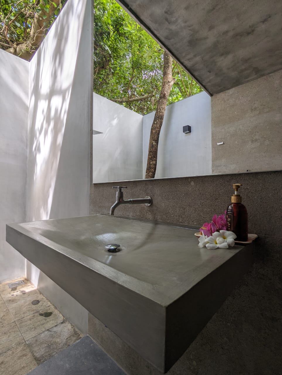 An outdoor bathroom space features a minimalist concrete sink with a single faucet. A small arrangement of purple flowers and a bottle of soap are placed beside the sink. Natural light filters through the surrounding greenery, creating a serene atmosphere.