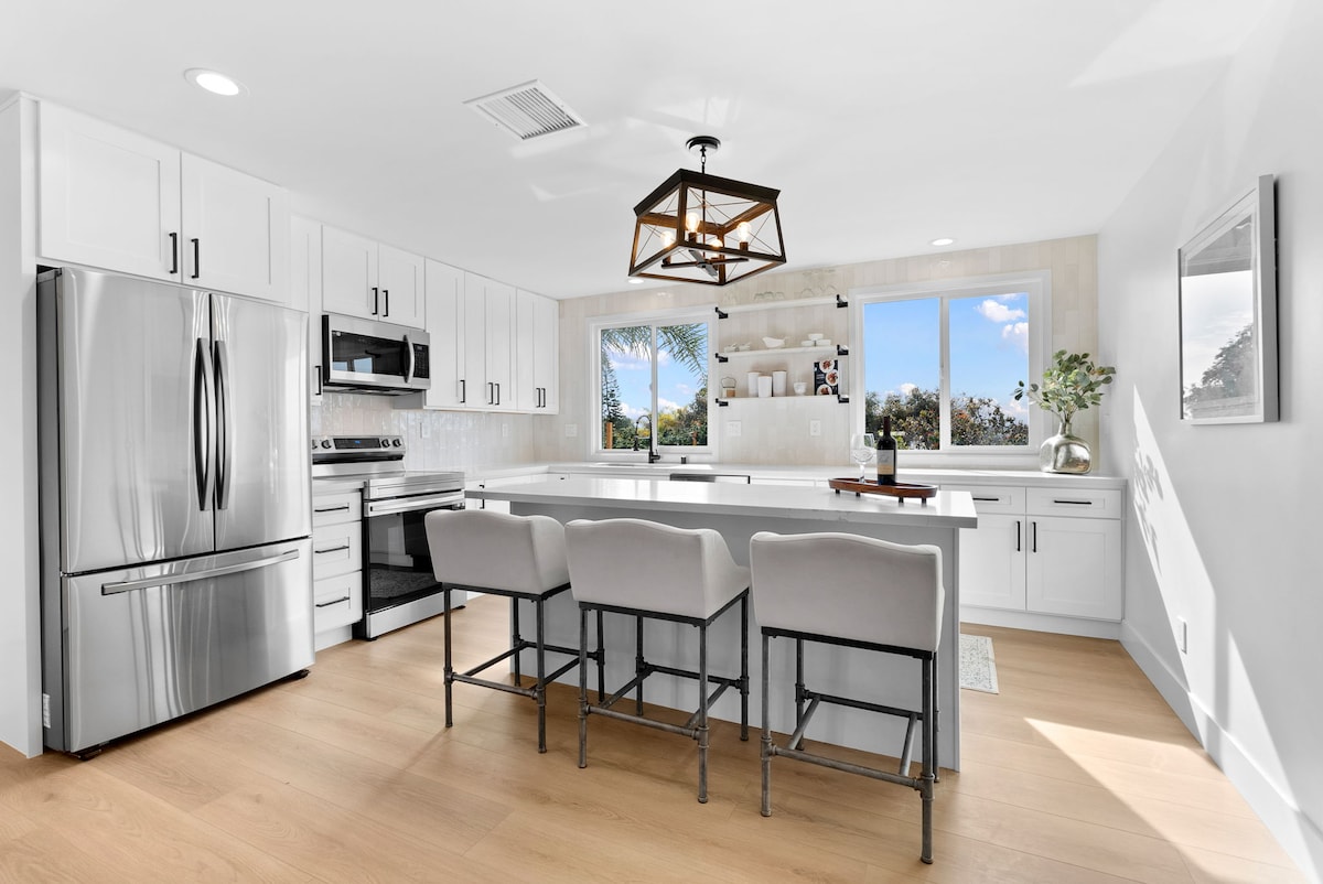 A modern kitchen is showcased with white cabinetry and stainless steel appliances. A spacious island with seating for three is situated beneath a decorative light fixture. Large windows offer views of the outdoors, allowing natural light to fill the room.