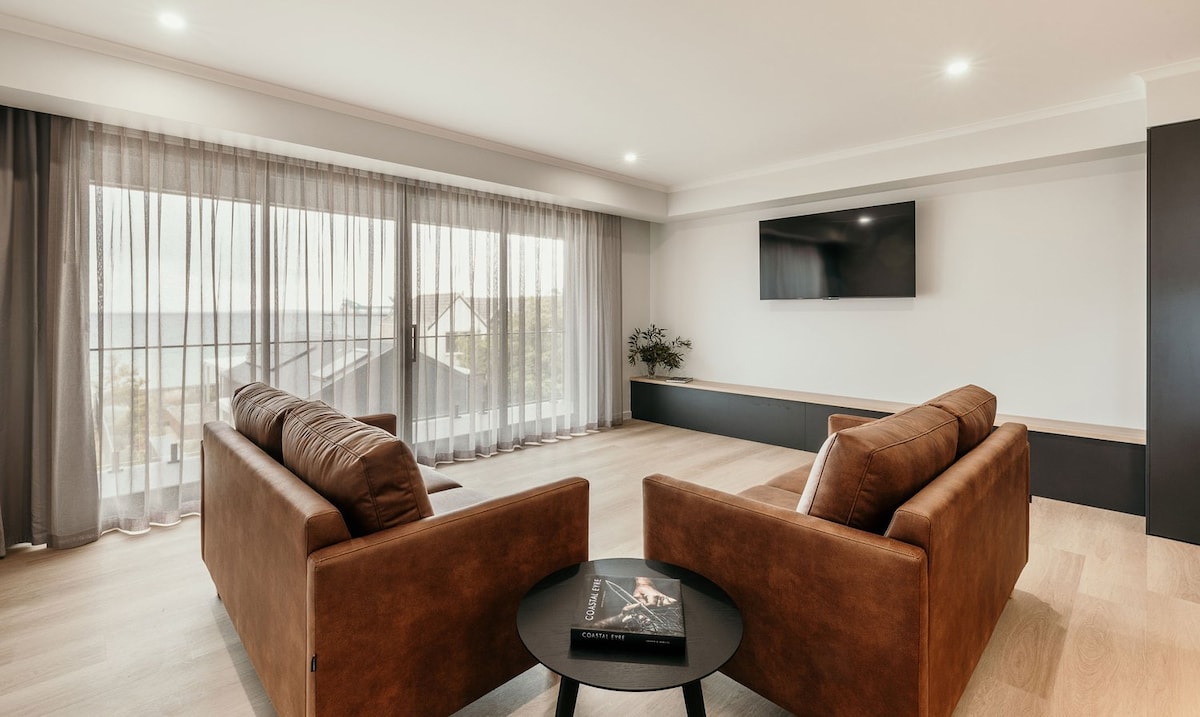 A spacious living area features two comfortable brown sofas facing a sleek flat-screen TV. Large glass sliding doors allow natural light and offer views of the outdoors. Sheer curtains gently diffuse the light, enhancing the openness of the space.