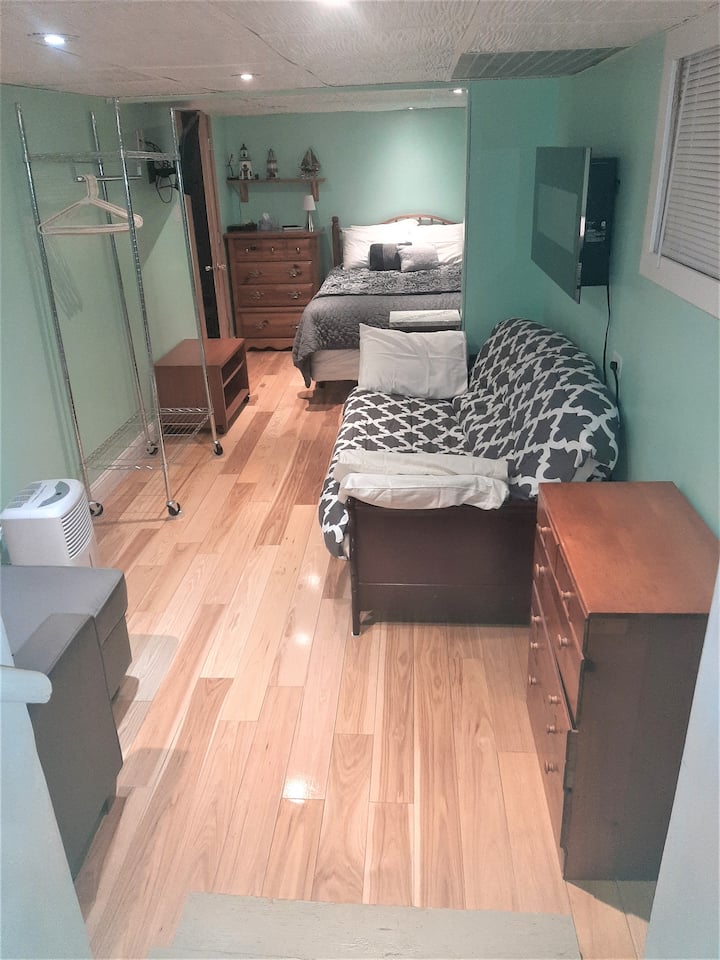 Spálňa na nižšej úrovni s manželskou posteľou v zadnej časti, futónom a stoličkou s pohovkou. Kúpeľňa sa nachádza vo vnútri otvorených dverí vľavo v zadnej časti obrázka. V tomto priestore spálne bola práve nainštalovaná podlaha z tvrdého dreva Hickory!