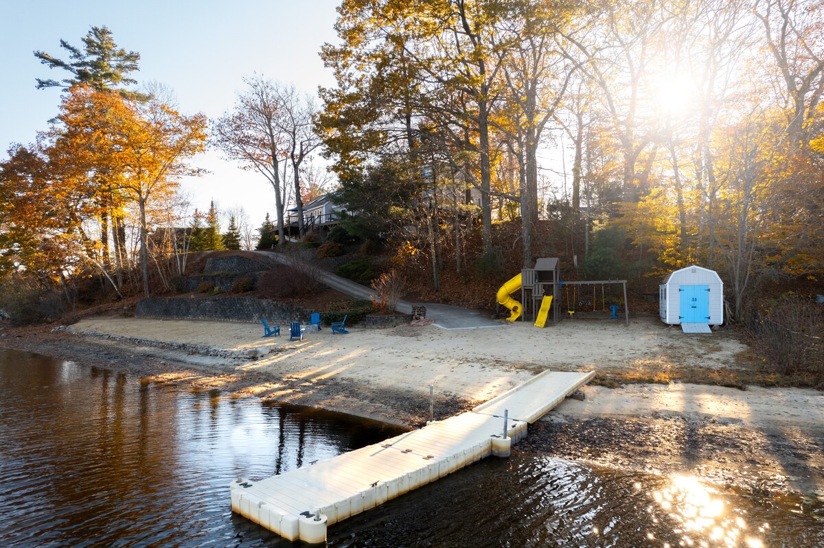 A sandy beach area extends along the water's edge, featuring a wooden dock and a playground with a yellow slide. Adirondack chairs are positioned nearby, surrounded by trees reflecting autumn colors, while sunlight filters through the foliage.