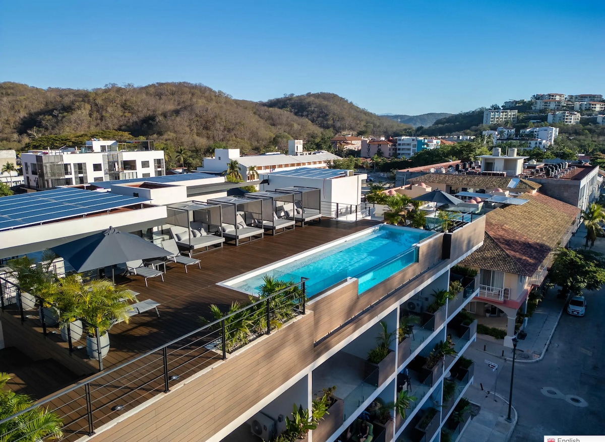 A rooftop patio is displayed, featuring an infinity pool surrounded by lounge chairs and shaded umbrellas. The area is bordered by tropical plants, providing a serene retreat. A panoramic view of the surrounding scenery, including buildings and hills, adds to the peaceful ambiance.