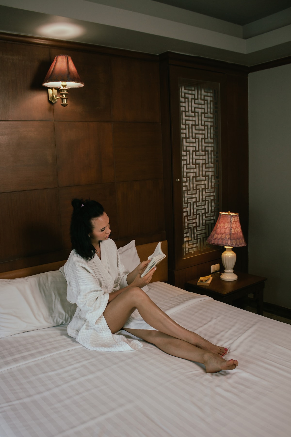 A spacious bed is draped with light-coloured linens, complemented by a wooden headboard featuring decorative panels. A bedside table holds a lamp with a warm glow. A guest is seated comfortably, reading a book while dressed in a robe, creating a relaxed atmosphere.
