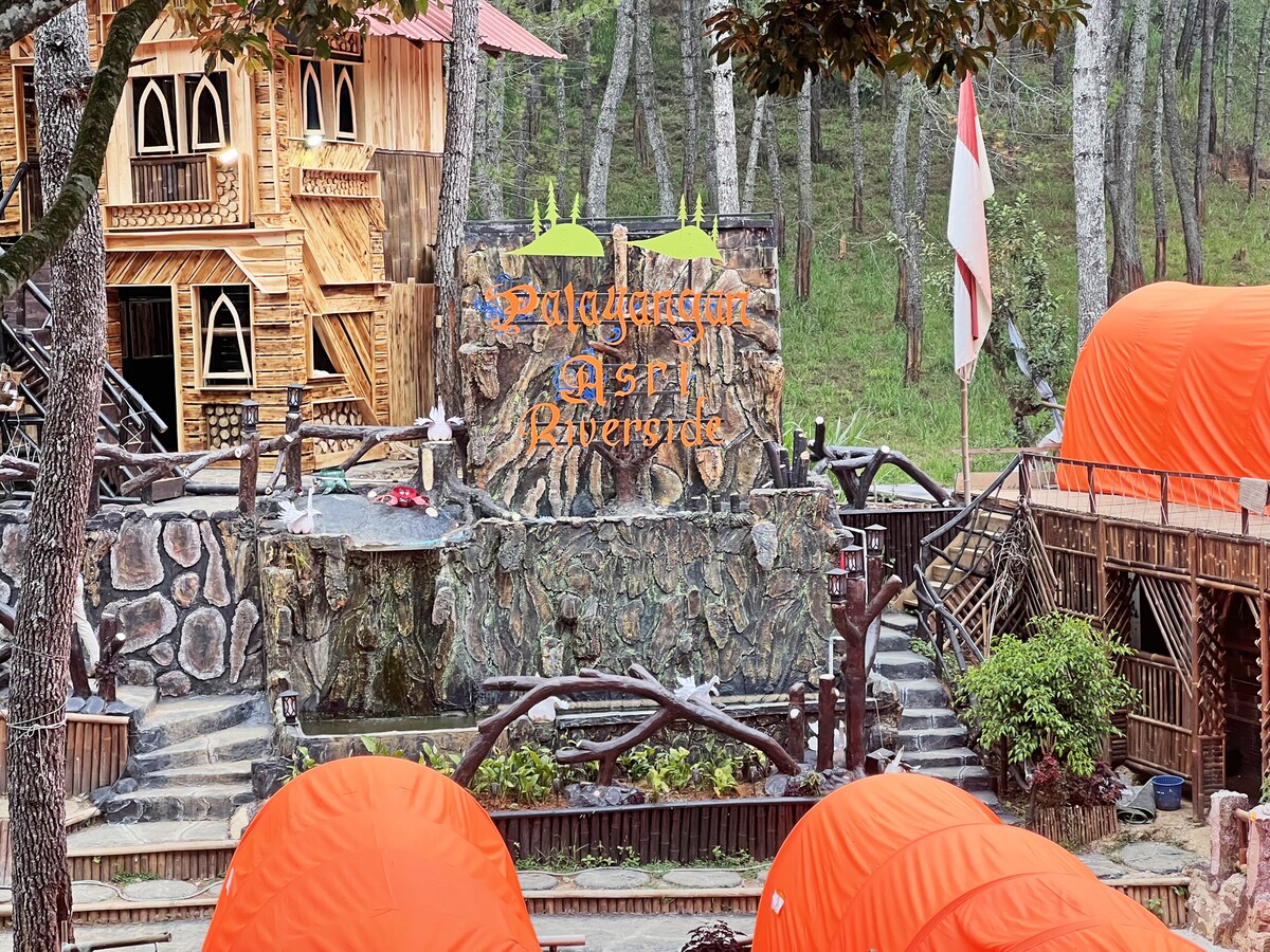 A rustic stone entrance sign is prominently displayed, featuring the name 'Palayangan Asri Riverside' in vibrant orange lettering. Surrounding the sign are natural stone pathways, greenery, and two orange glamping tents, set against a backdrop of tall trees in a serene woodland environment.