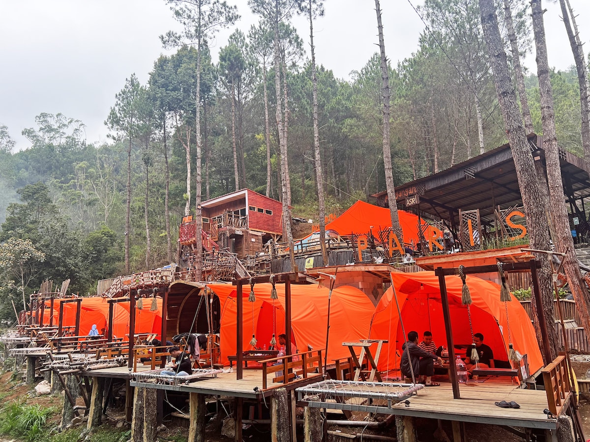 A series of vibrant orange tents are arranged on a wooden platform among tall trees. The natural setting is complemented by rustic wooden tables and chairs. In the background, a multi-level structure features a red facade and visible signage, adding to the inviting ambiance.