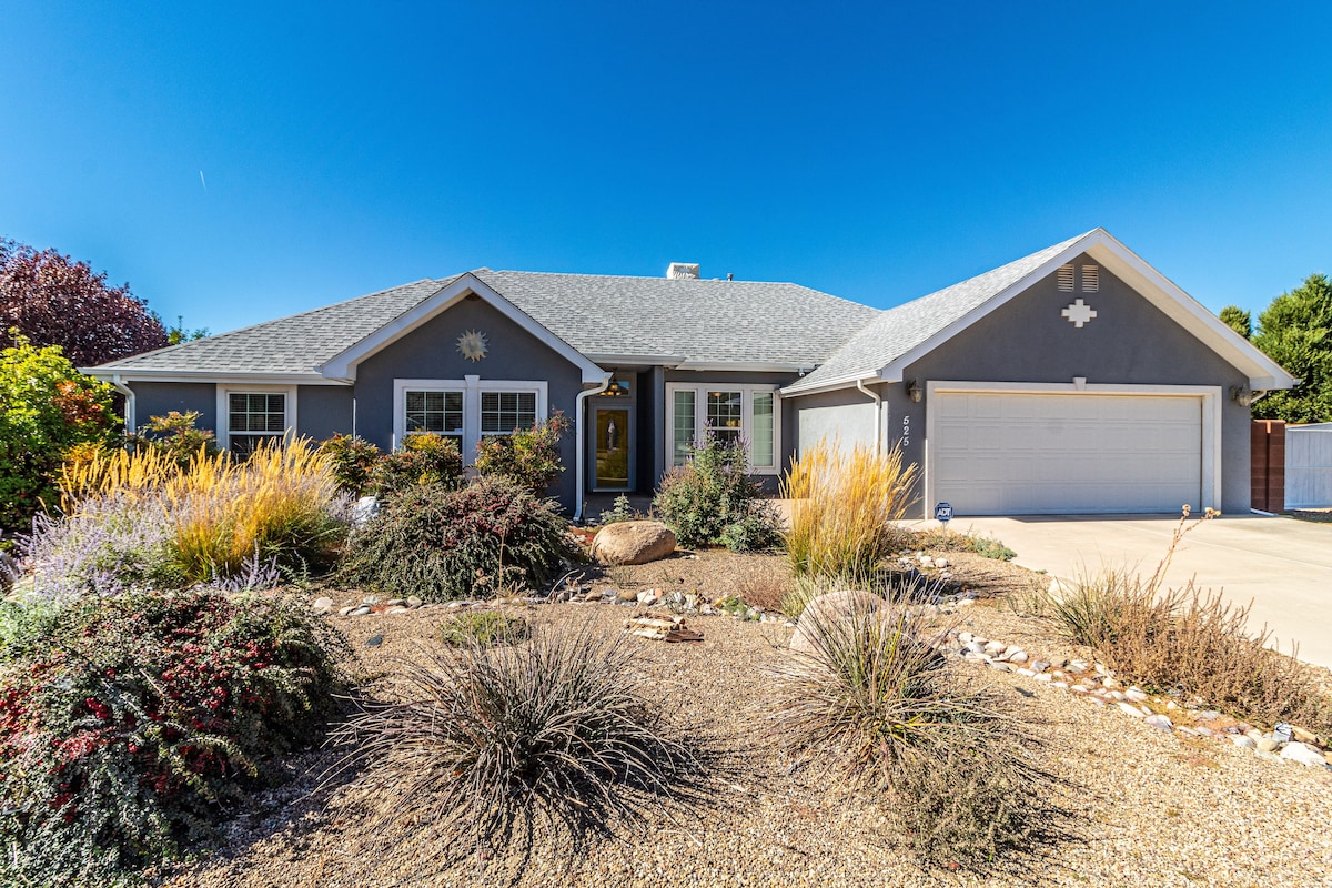 A single-story home features a well-maintained front yard with a variety of colorful shrubs and ornamental grasses. The house is accented by a charming entrance and a clean driveway leading to a spacious garage. Clear blue skies complement the inviting exterior.