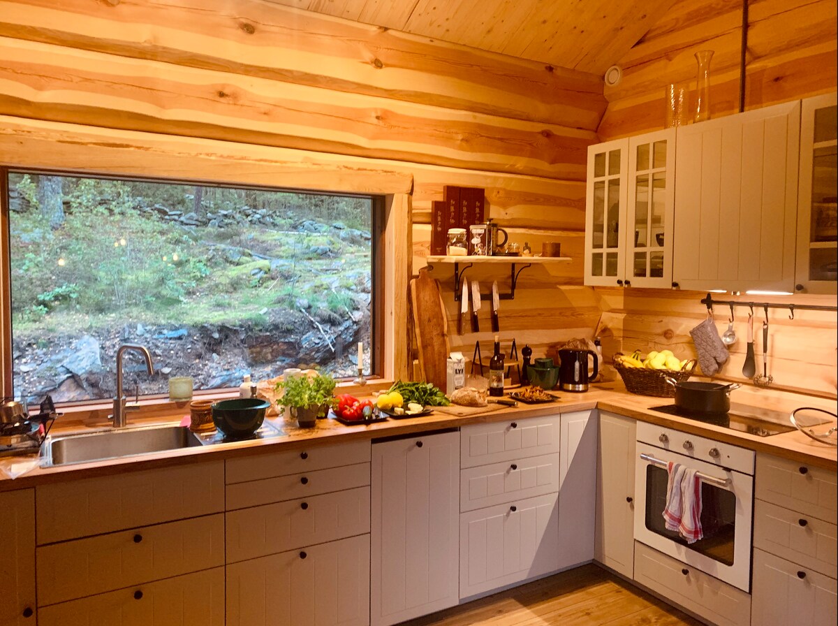 A modern kitchen features ample counter space with a variety of fresh vegetables and fruits displayed. A large window offers a view of the surrounding forest, while cabinetry and appliances complete the functional layout.