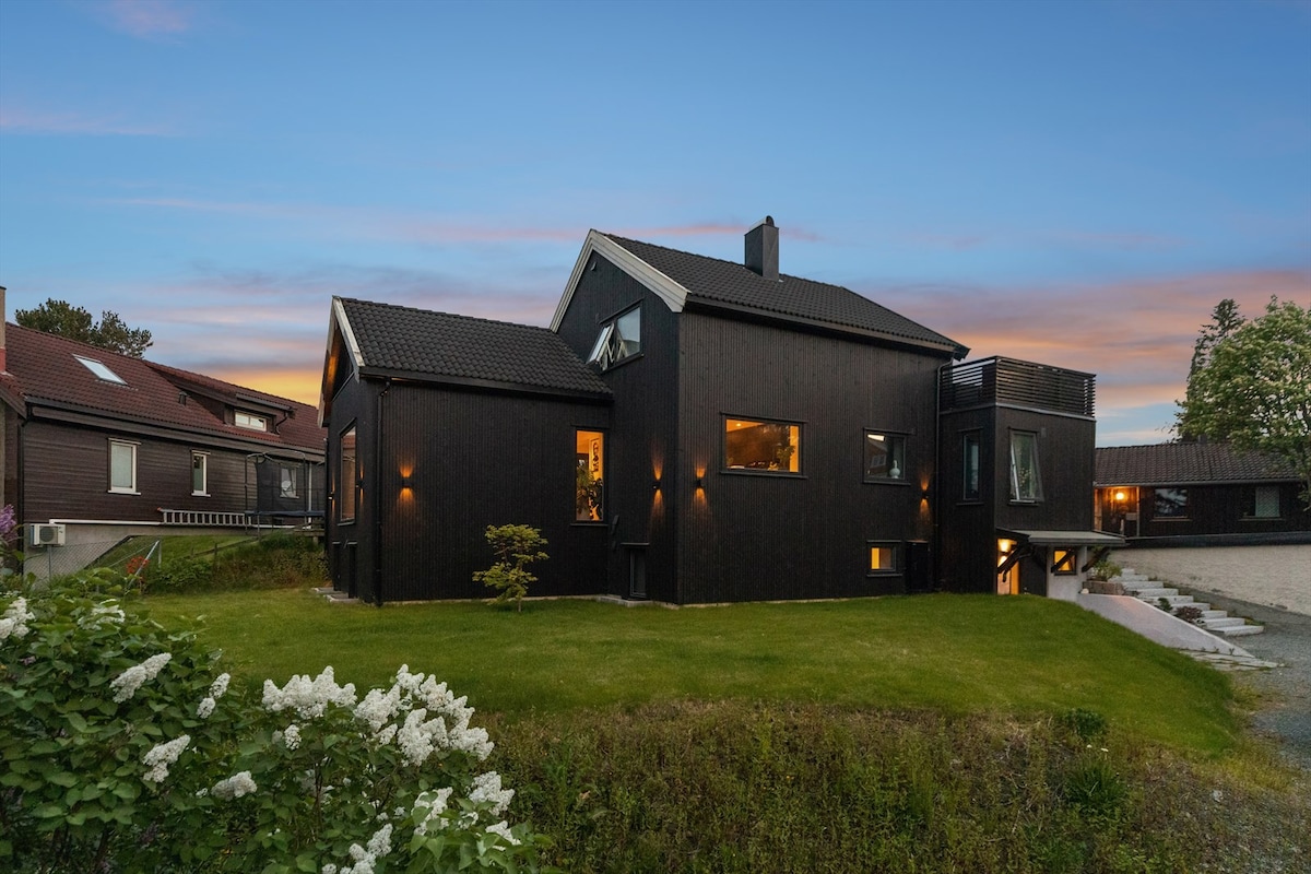 The modern, dark-hued house is framed by manicured green grass and white flowering shrubs. Large windows offer glimpses of the interior, while outdoor lighting enhances the evening ambiance. The building's unique architecture is complemented by a serene setting, with neighboring houses visible in the background.