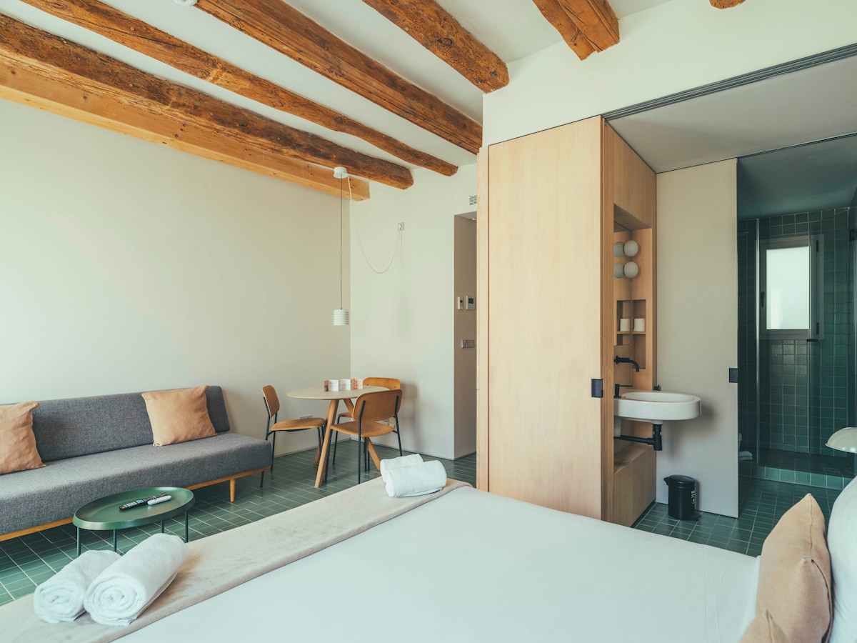 A spacious room features exposed wooden beams and offers a view of a cozy seating area with a sofa and dining table. A queen size bed is positioned nearby, and a bathroom is visible, showcasing a modern sink and shower area.