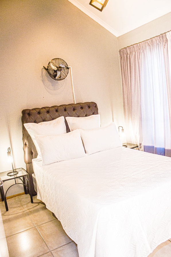 A cozy sleeping area features a tufted headboard with white bedding arranged neatly on a spacious bed. Soft natural light filters through sheer curtains, illuminating bedside tables with lamps on either side.